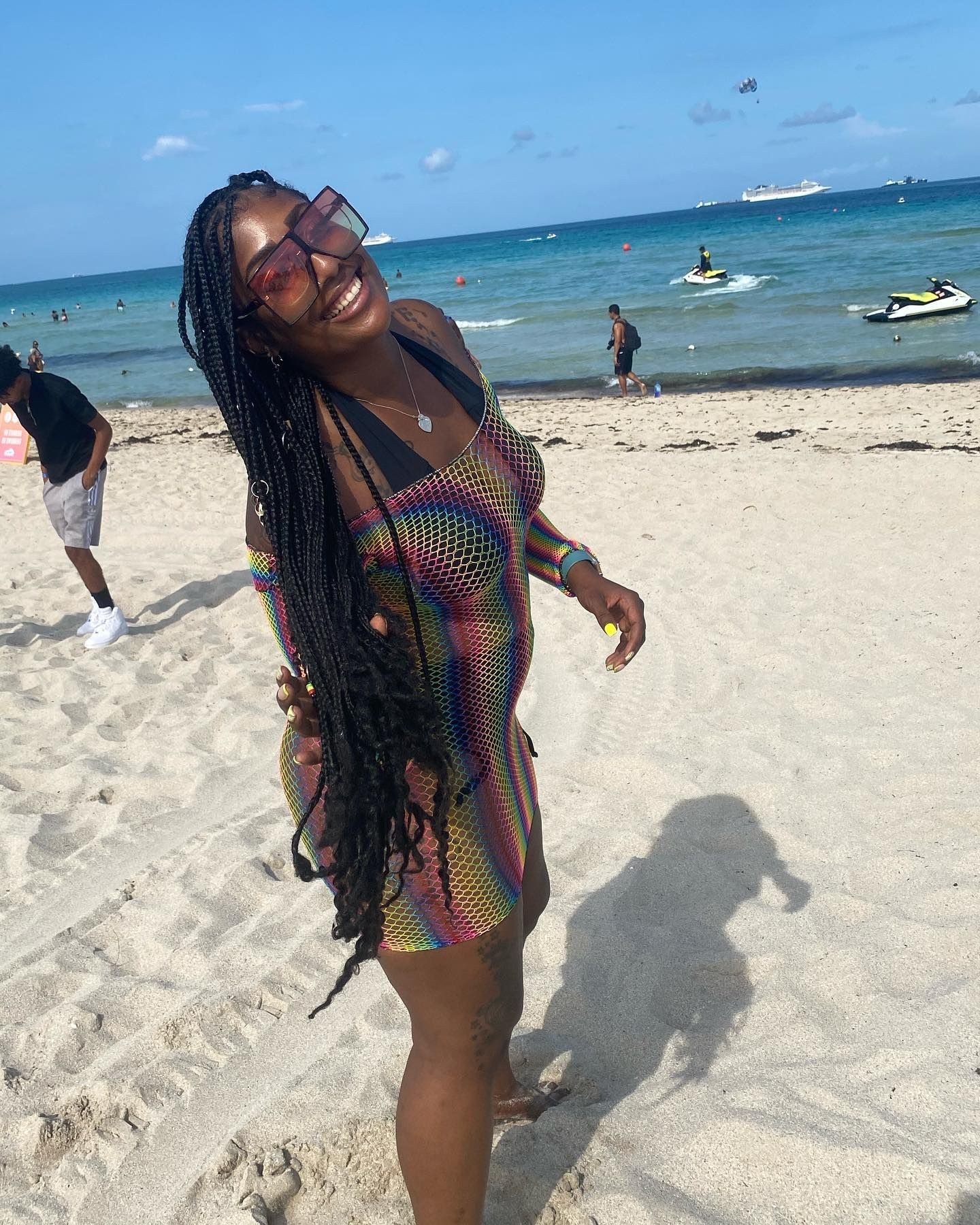 Woman in colorful dress smiling on a sandy beach with ocean and jet skis in the background.