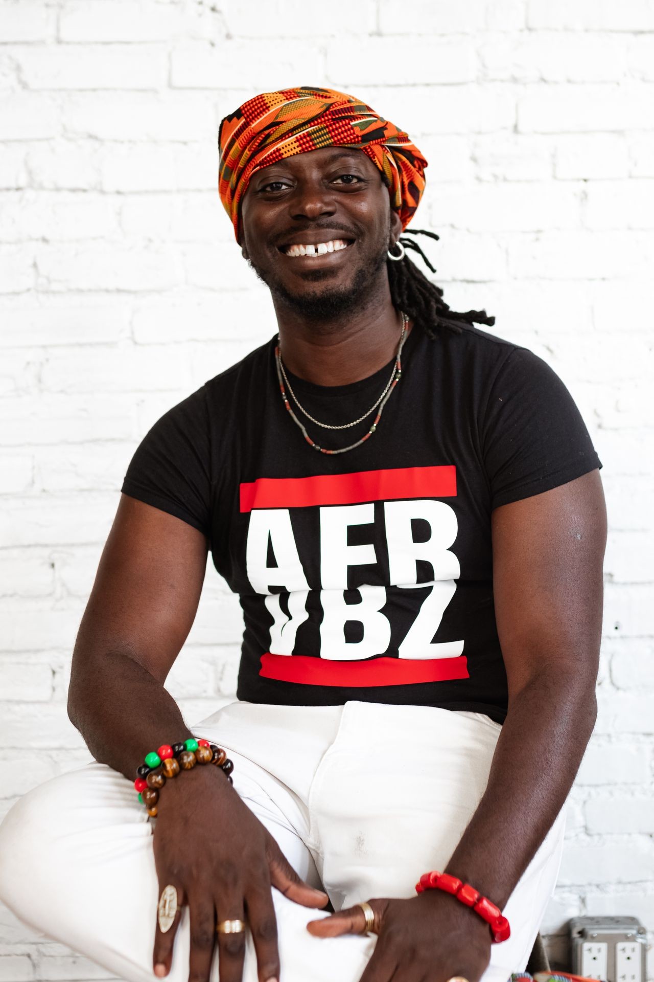 Smiling person wearing colorful headscarf and black T-shirt against white brick wall.