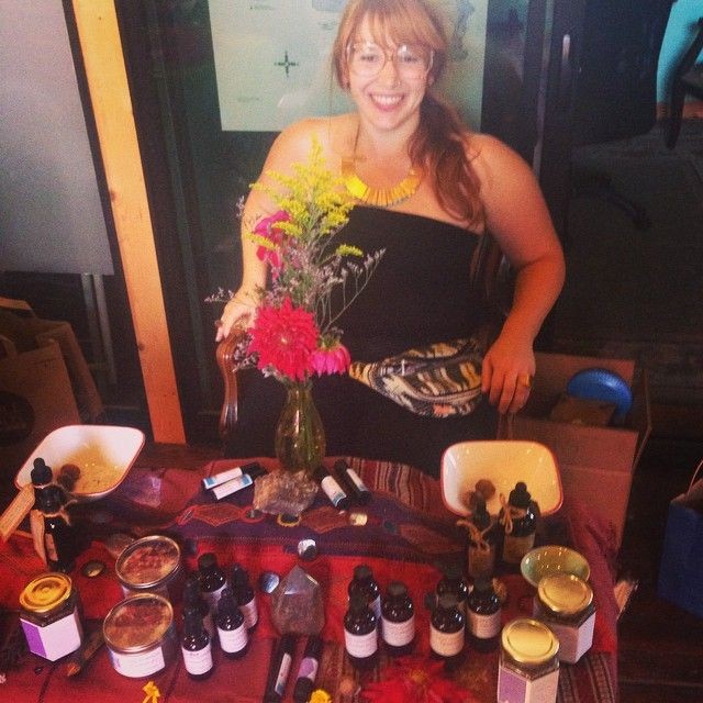 Smiling woman sitting behind a table with homemade products and a vase of flowers.